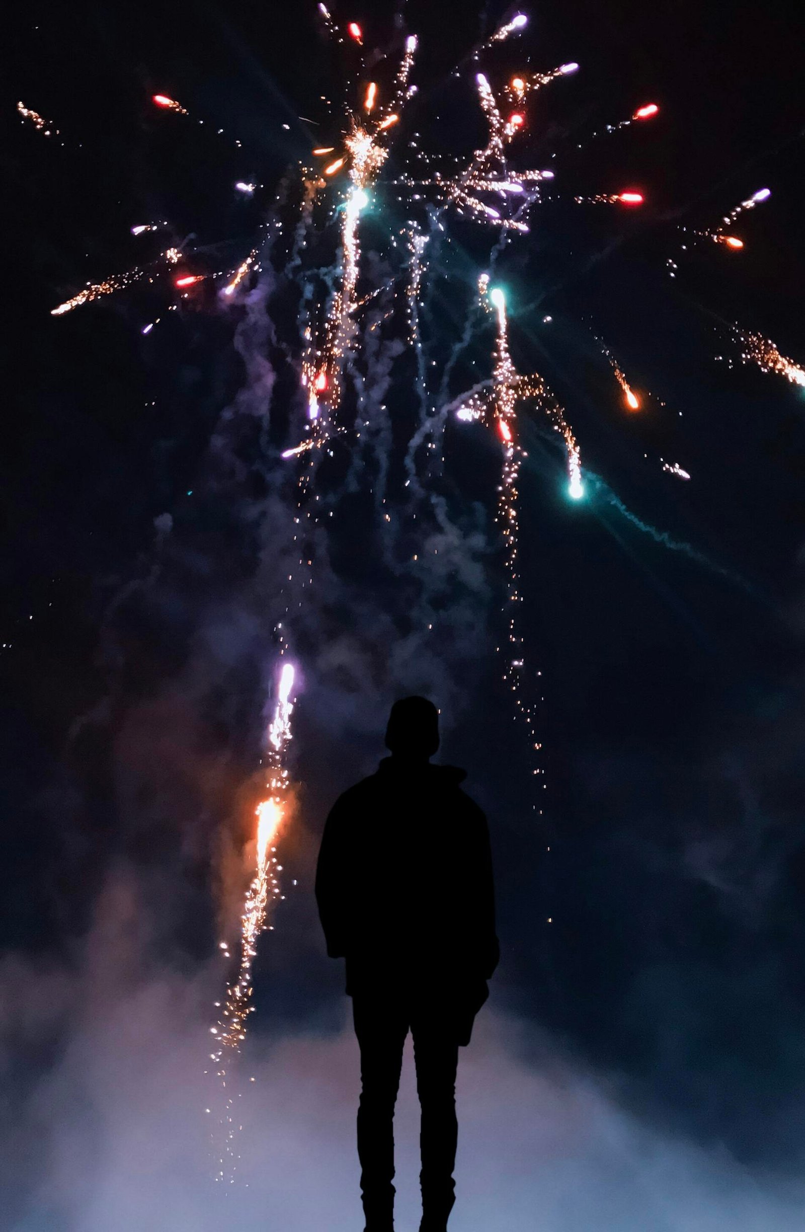 A person silhouetted against a colorful night sky with fireworks bursting overhead.