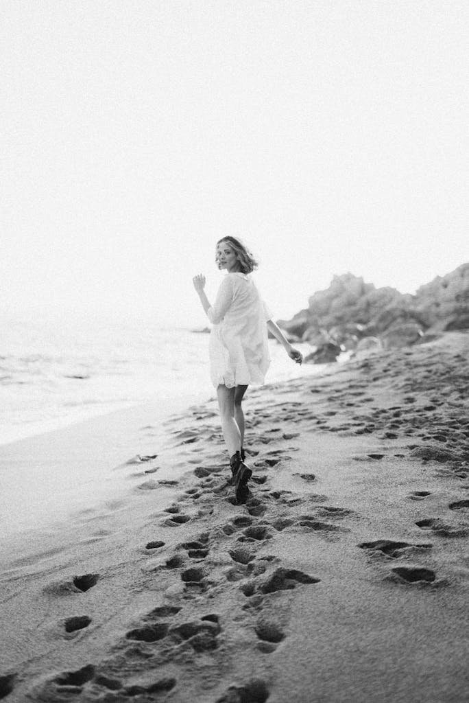 A woman in a white dress walks along a sandy beach, leaving footprints behind in a serene black and white scene.