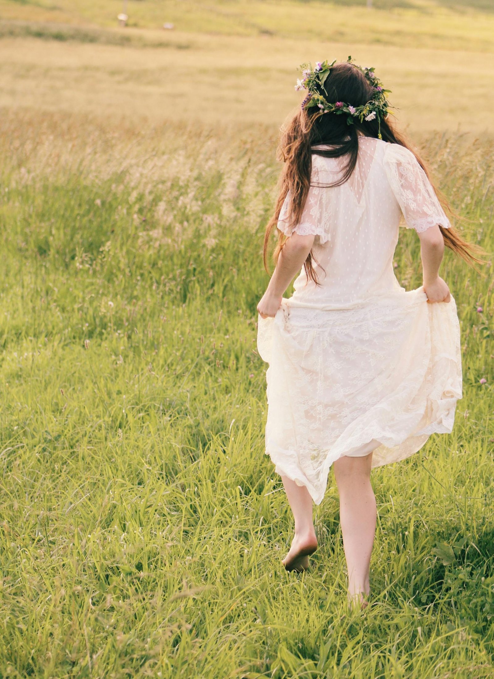 A young woman in a flowing white dress and flower crown walks barefoot in a sunlit field.