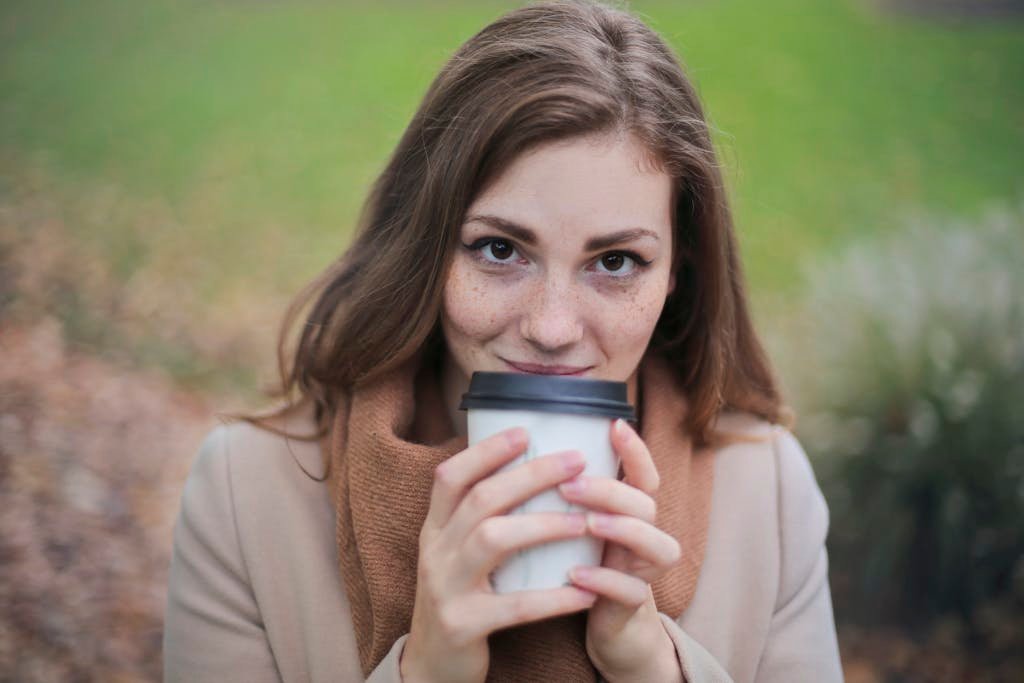 A young woman smiling while holding a coffee cup in a park during fall.