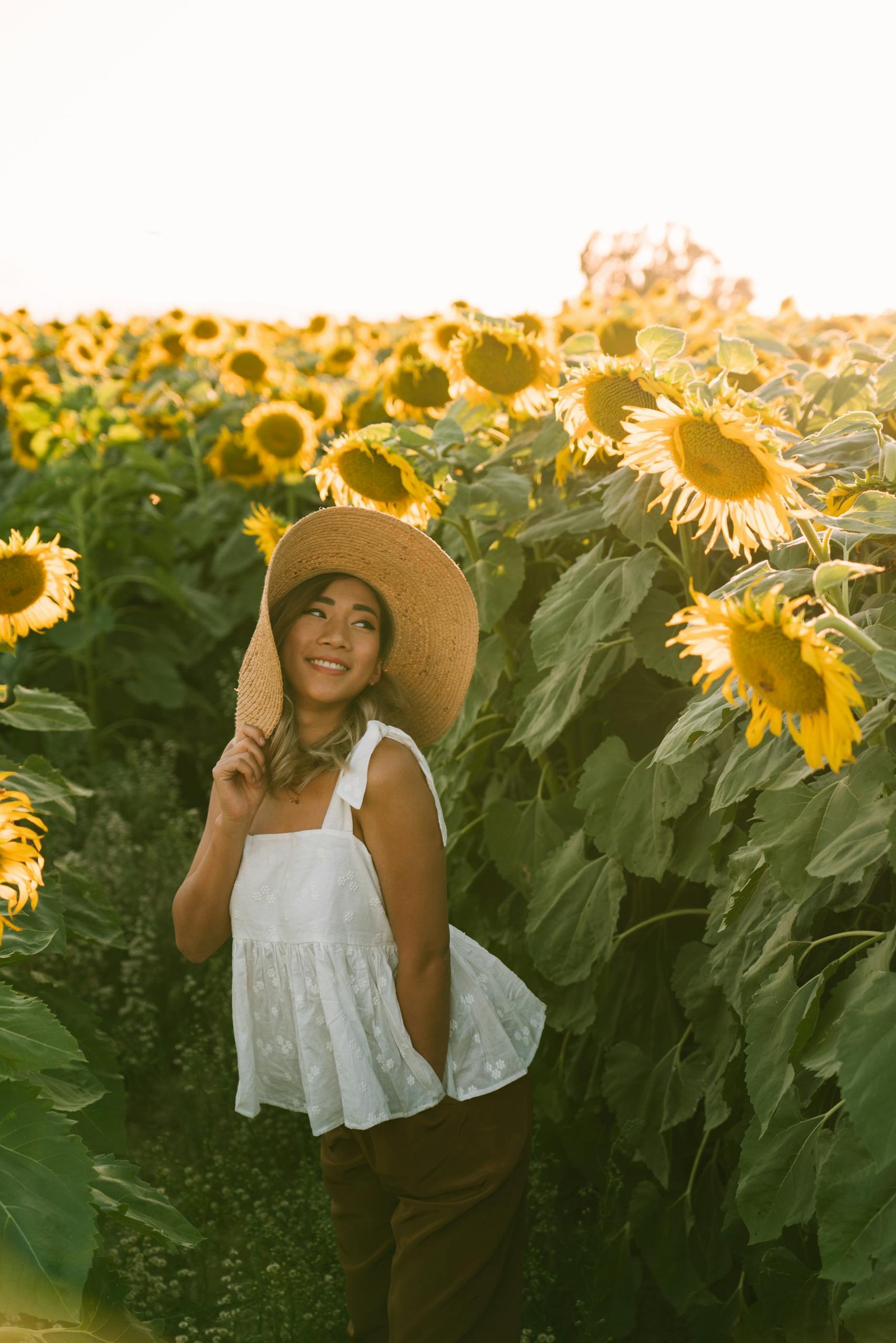 Asian woman in a sunflower field, smiling under a sun hat during a beautiful sunset.
