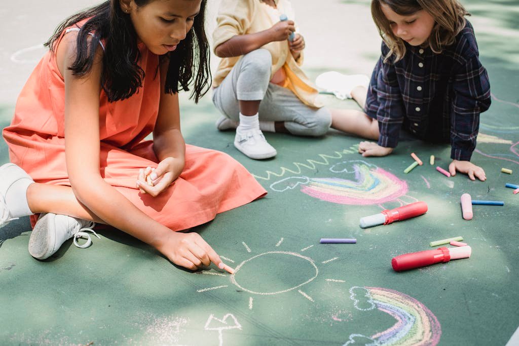 Three children drawing colorful chalk art on pavement, depicting rainbows and sunshine.