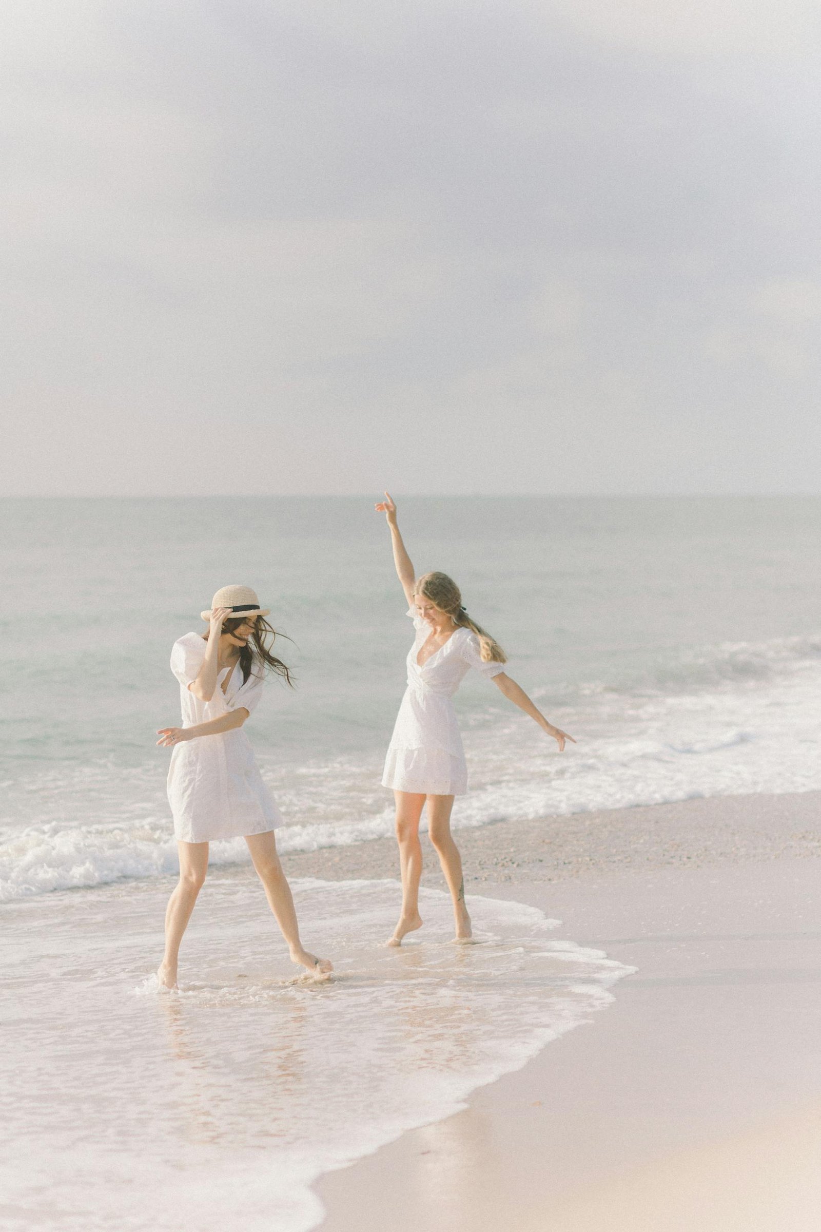 Two women in white dresses enjoying a carefree moment on a sunny beach by the sea.