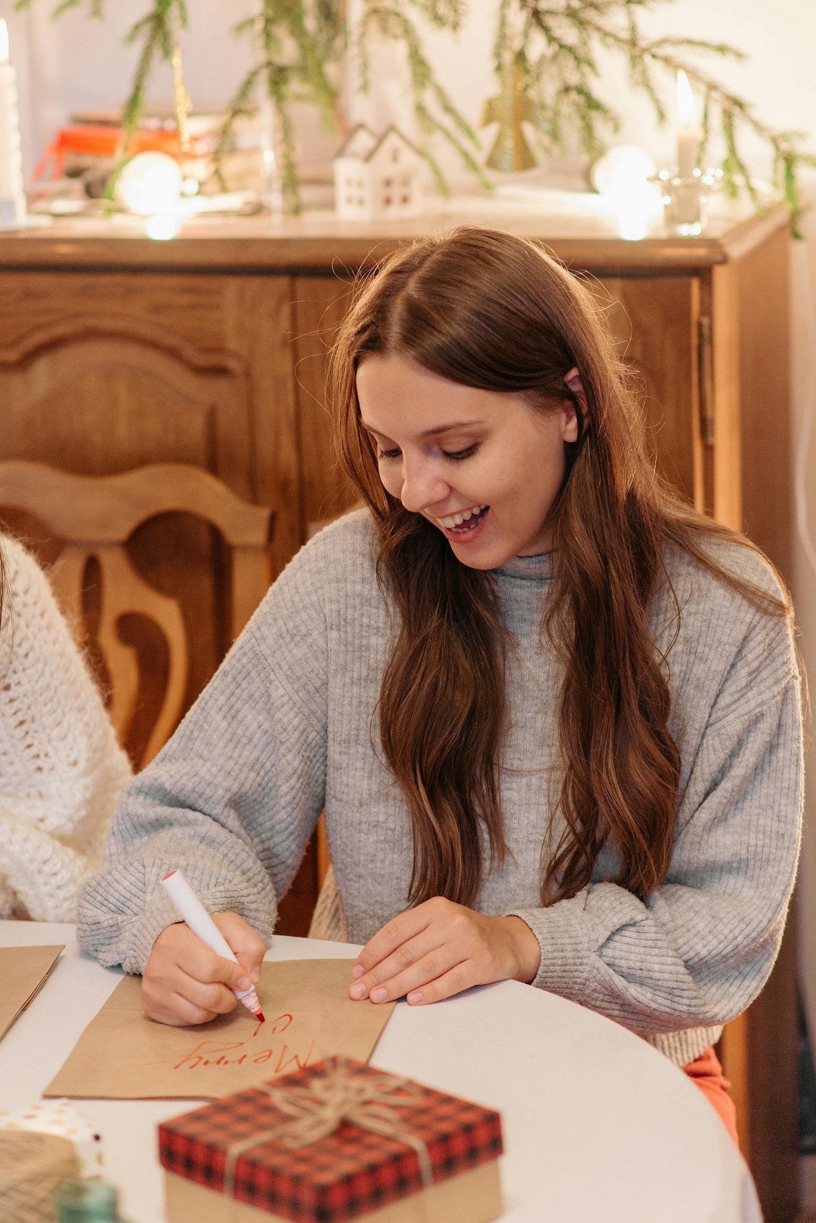 Happy woman in a gray sweater writing a holiday card at a festive table with gifts and decorations.